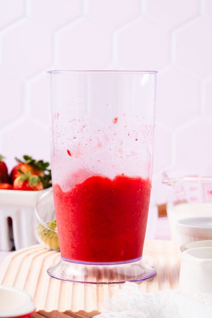 A clear blender cup filled with blended red strawberry puree on a light wooden surface, perfect as a base for your favorite Strawberry Matcha Recipe.