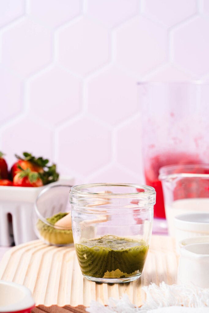 A glass jar with green liquid sits on a wooden surface, surrounded by strawberries and other ingredients&mdash;perfect for creating a refreshing Strawberry Matcha Recipe.