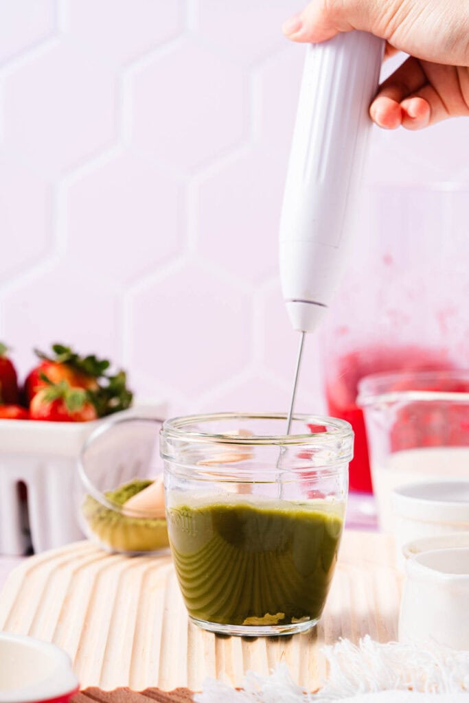 A hand froths green matcha in a glass jar with a handheld frother on a wooden surface, preparing to create a refreshing Strawberry Matcha Recipe.