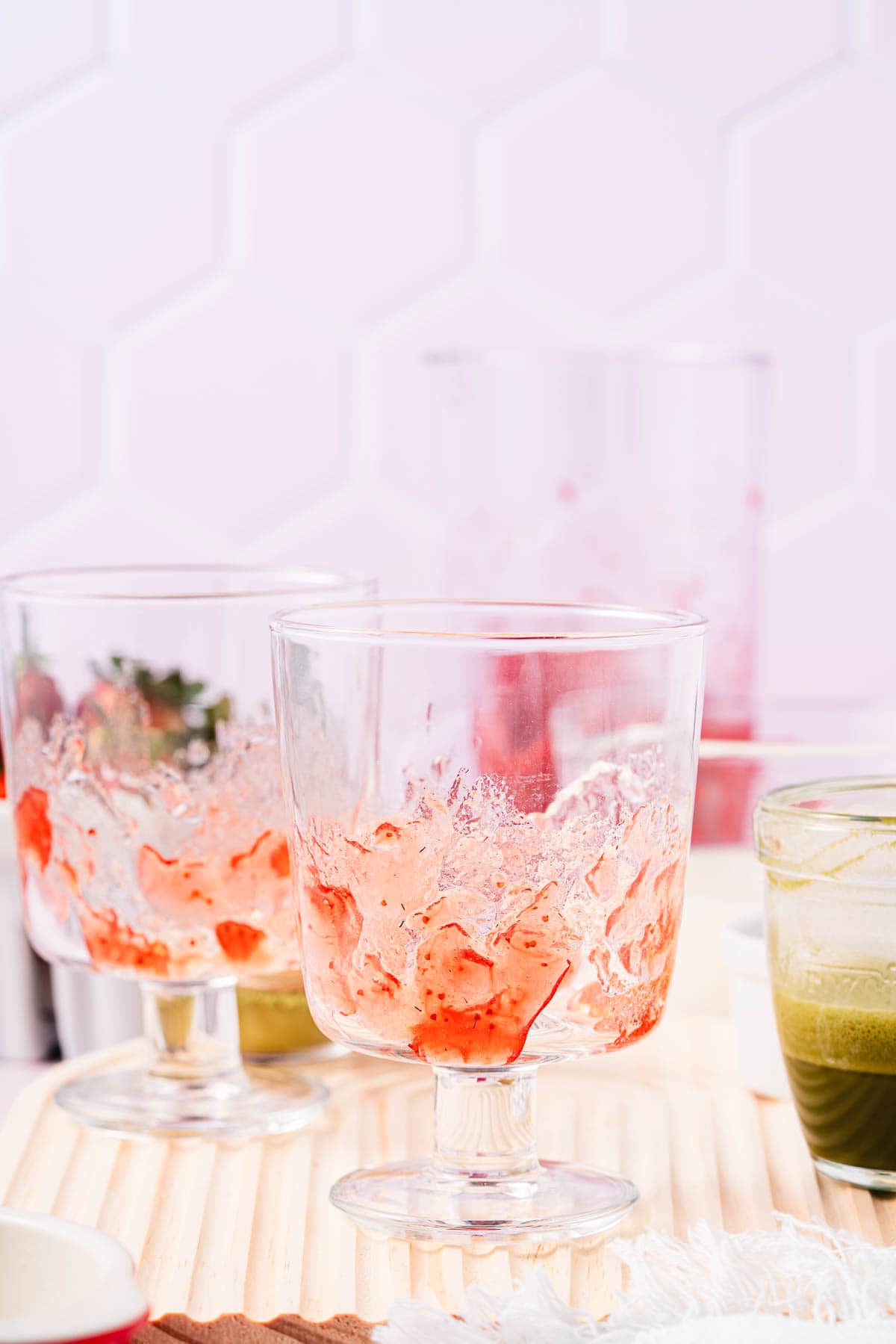 Two empty dessert glasses with strawberry remnants sit beside a glass of green liquid, hinting at a recently enjoyed Strawberry Matcha Recipe, all on a light wooden surface.