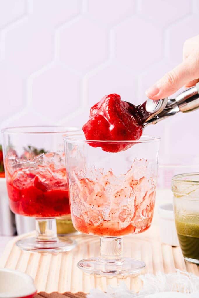 A hand scoops bright red strawberry sorbet into a glass, with another glass of the fruity recipe nearby on a wooden surface.