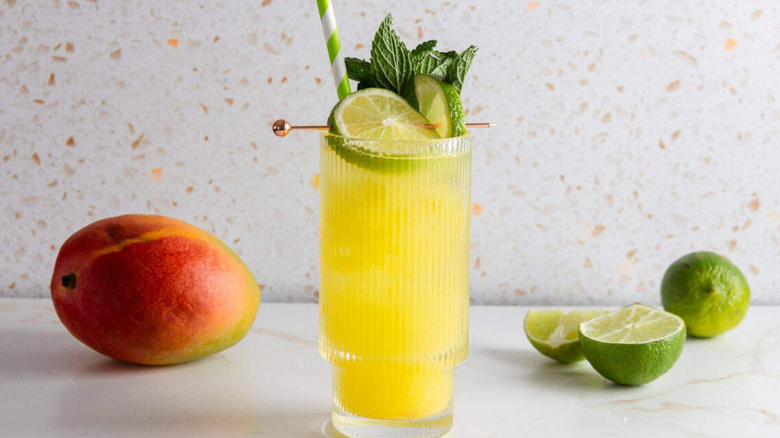 A mango mocktail with lime slices, mint, and a striped straw, beside a whole mango and limes.