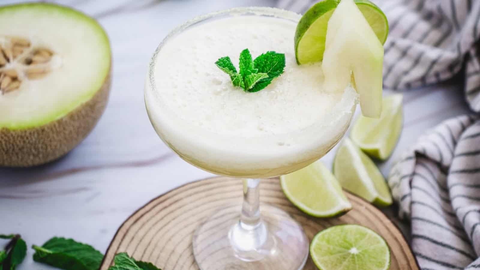 A frozen melon cocktail garnished with lime, melon slice, and mint leaves on a wooden board.
