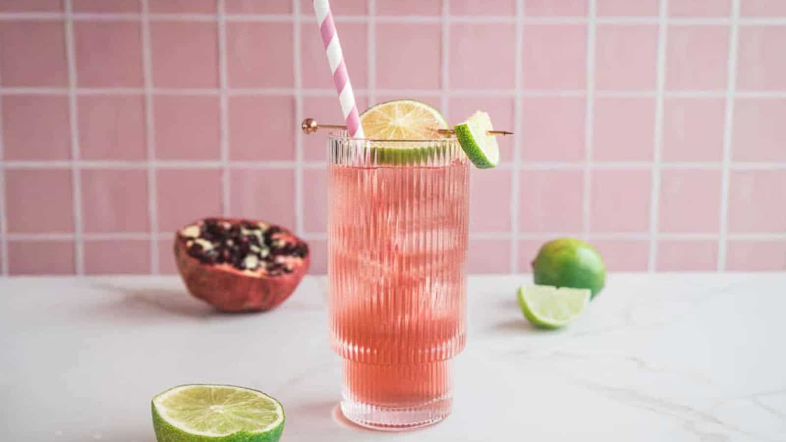 A pink drink with ice, lime slices, and a striped straw in a glass, with fruit on a white surface.