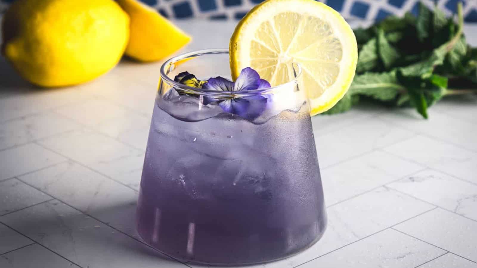 A glass of purple iced drink garnished with a lemon slice and a flower, with lemons and mint in the background.