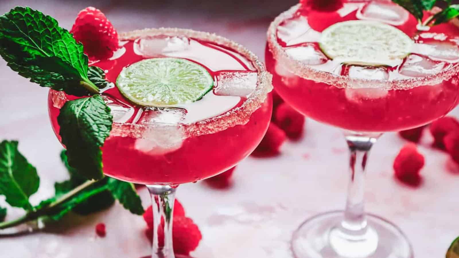 Two glasses of pink cocktail with ice, lime slices, sugared rims, mint, and raspberries as garnish.