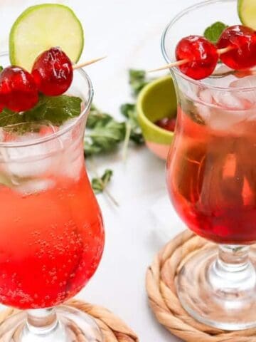 Two red iced drinks in curved glasses, garnished with cherries, lime slices, and mint, on woven coasters.