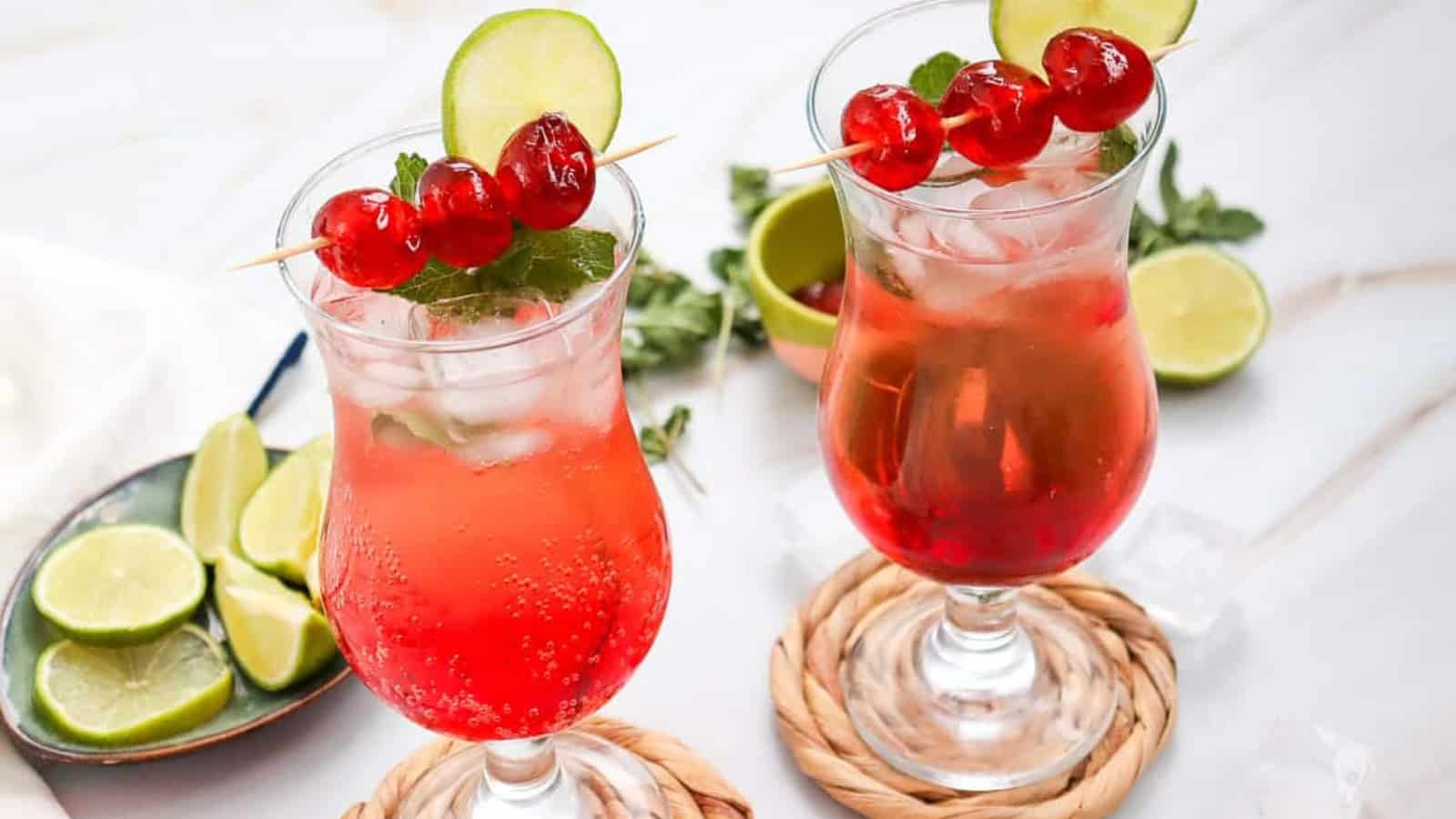 Two red cocktails with ice, garnished with lime, cherries, and mint, on woven coasters with lime slices nearby.