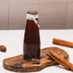 A glass bottle of dark syrup on a wooden board with cinnamon sticks and nutmeg nearby.