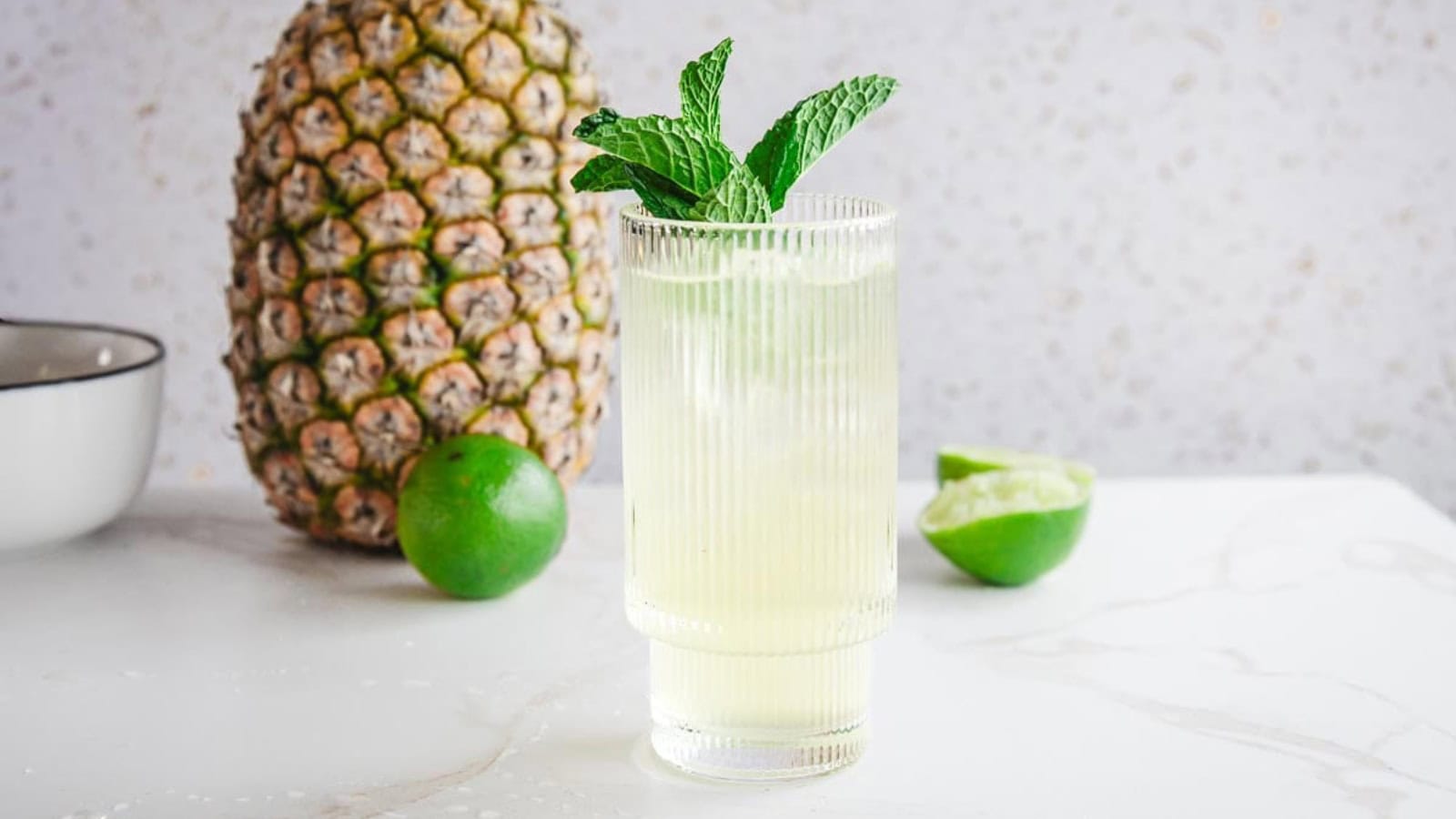 A glass of light yellow drink with mint, next to a whole pineapple and limes on a white surface.