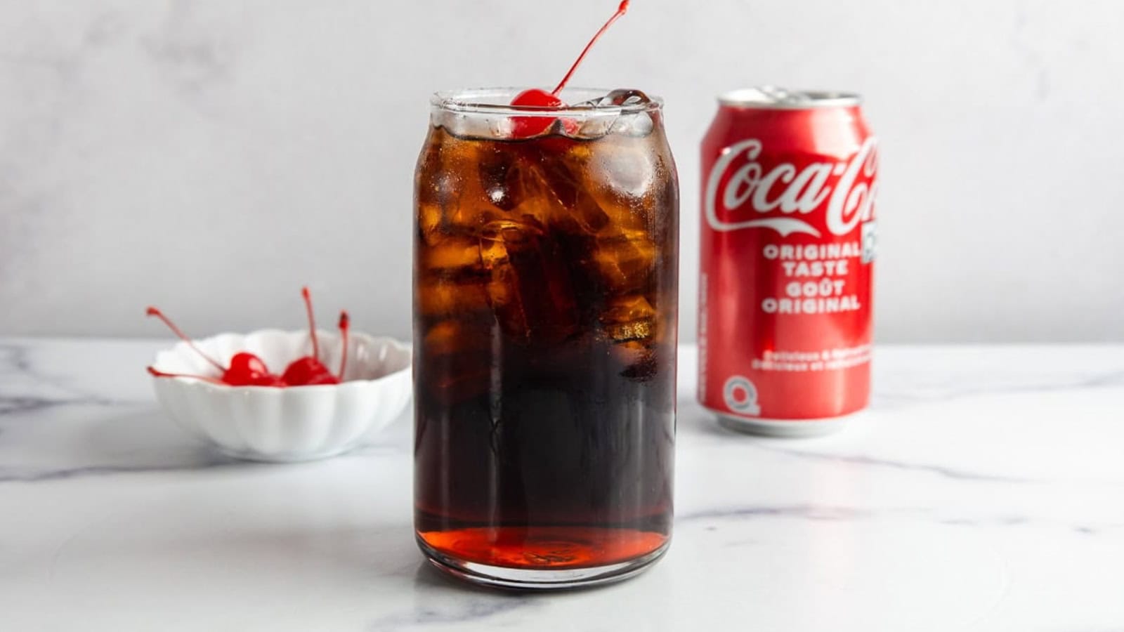 A glass of cola with ice and a cherry, next to a can of Coca-Cola and a bowl of cherries.