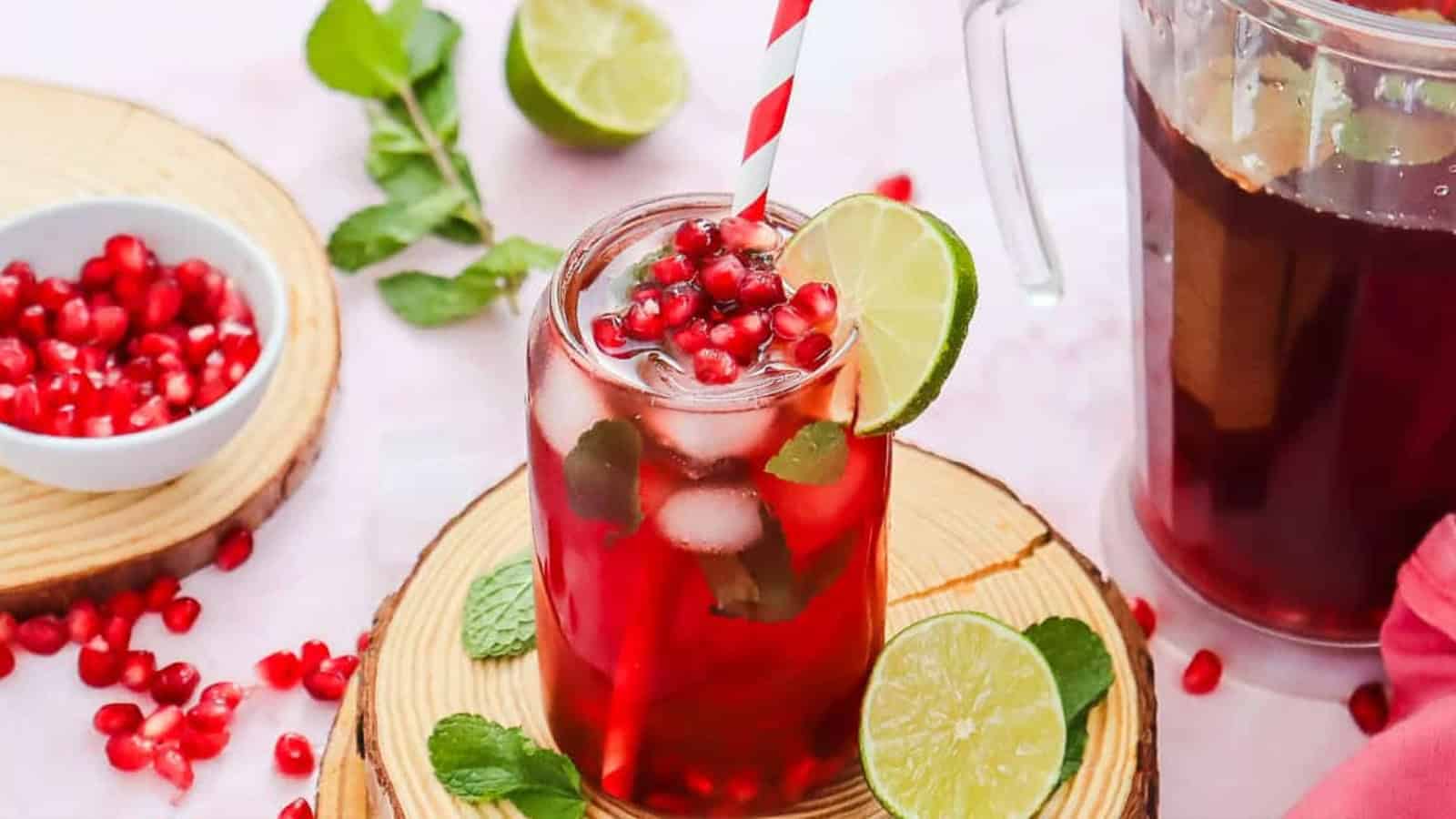 A glass of pomegranate drink with ice, mint, lime slice, and a straw, surrounded by pomegranate seeds.
