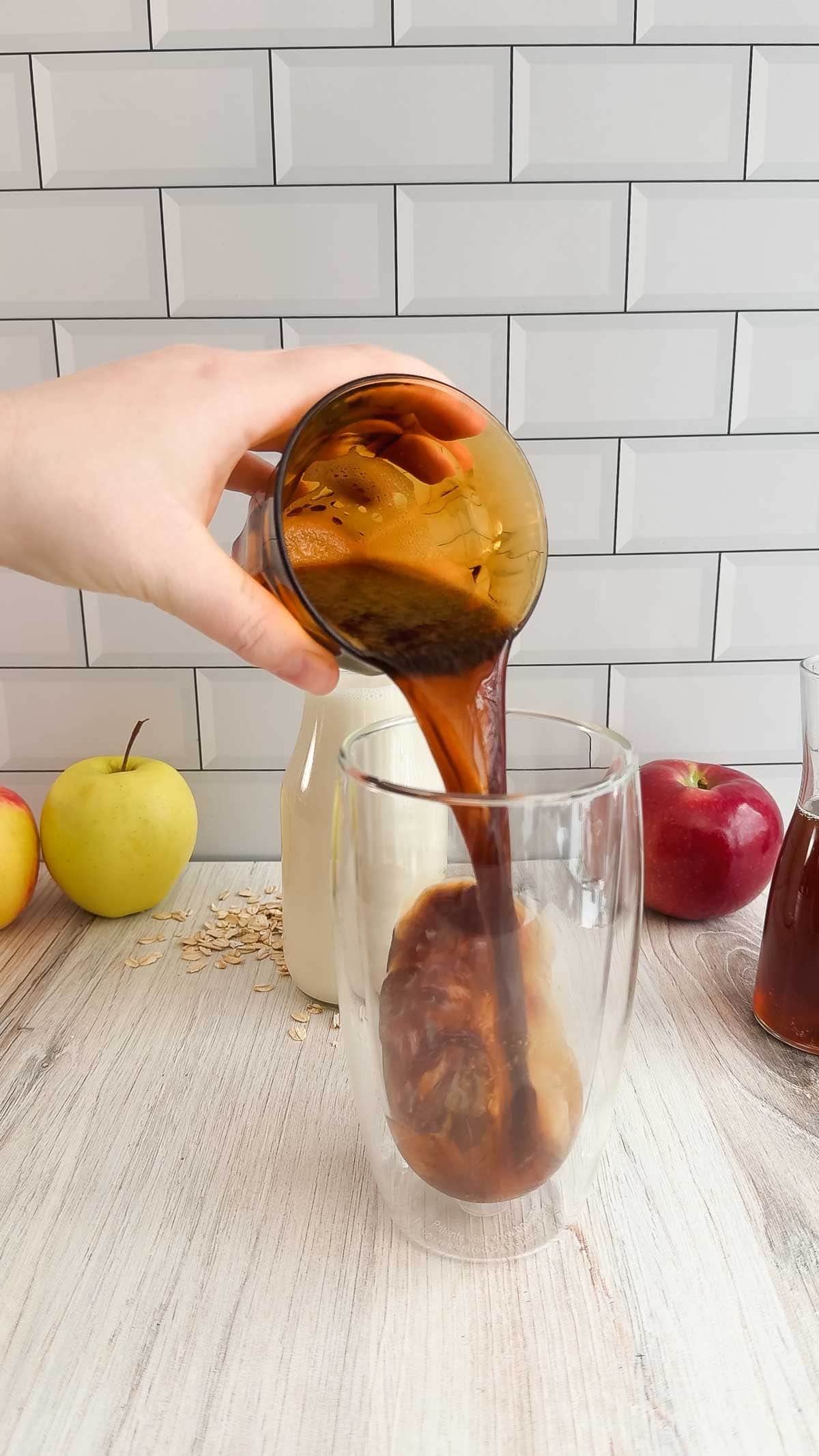 A hand pours a Starbucks copycat apple crisp latte into a glass on a wooden table, with apples and bottles in the background.