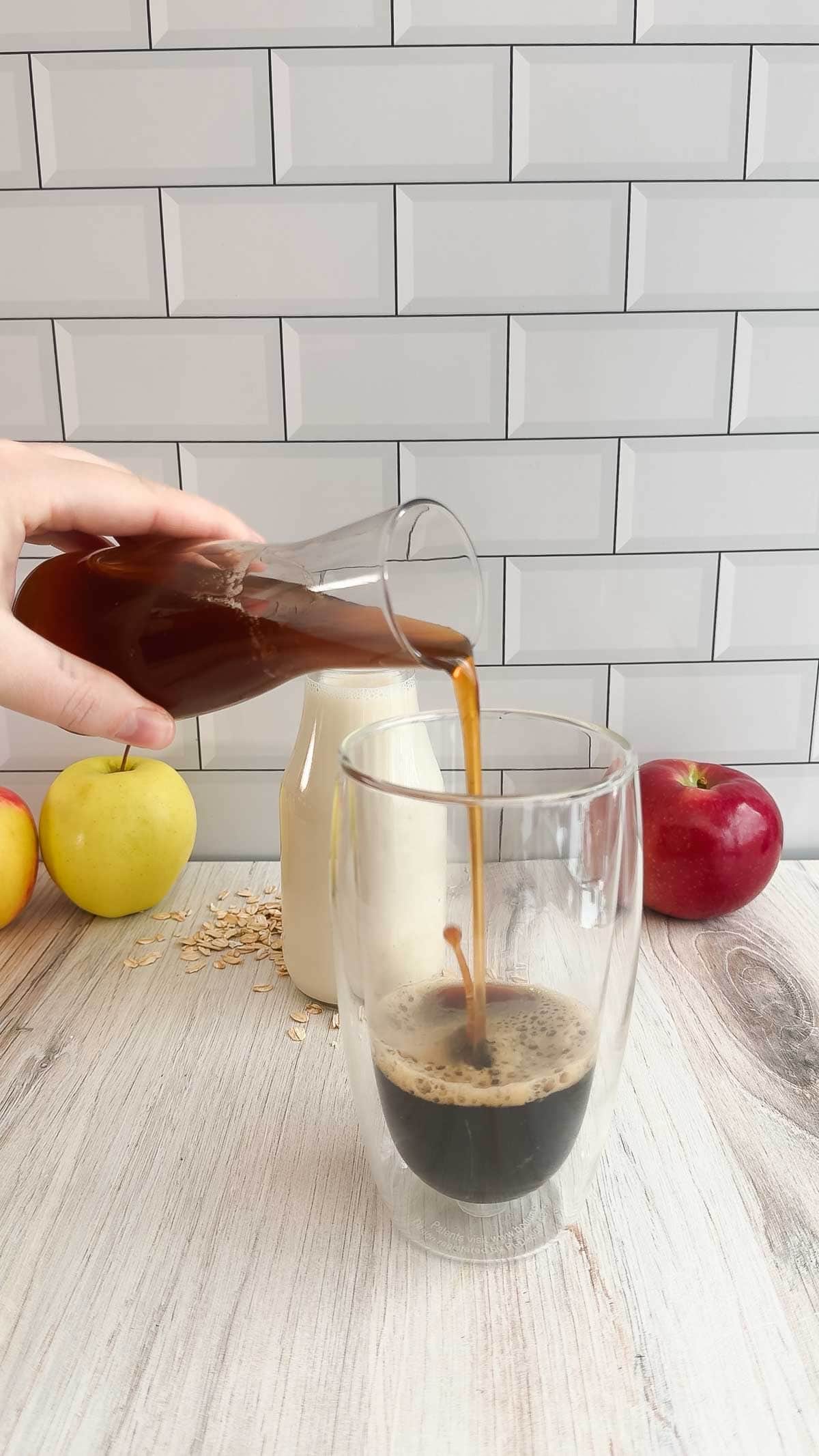 Hand pouring coffee into a glass for an Apple Crisp Latte with apples, oats, and a bottle on a wooden table in the background&mdash;a perfect Starbucks Copycat Oat Milk Latte experience.