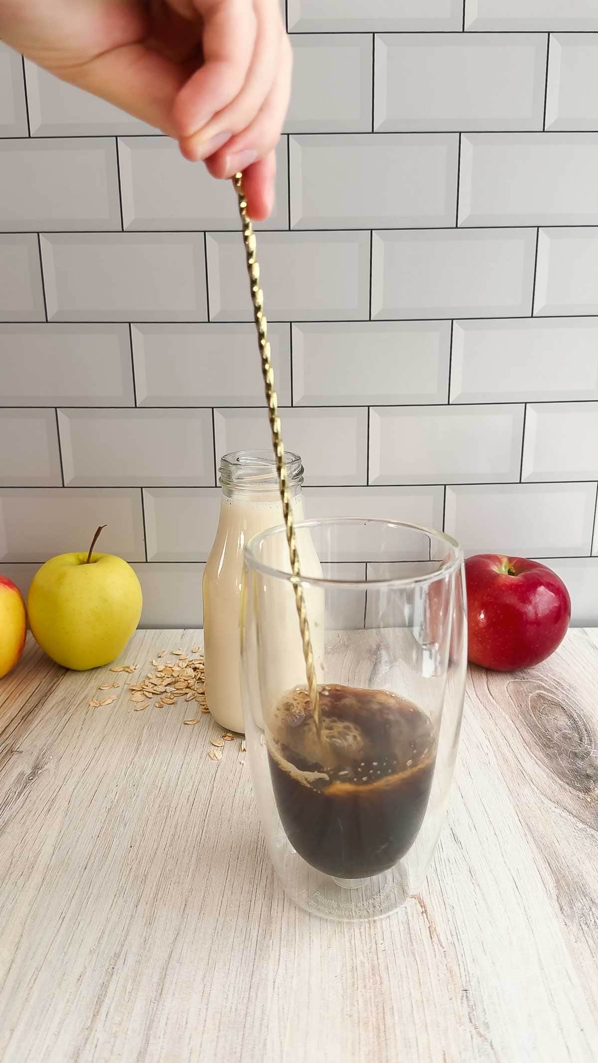 A hand stirs iced Apple Crisp Oat Milk Latte in a glass with a gold spoon; apples and a milk bottle add to the Starbucks Copycat experience in the background.