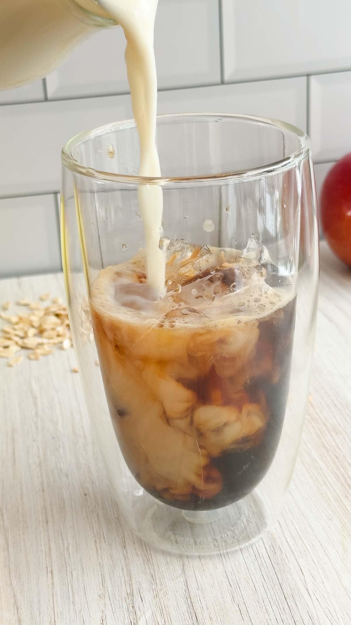 Milk being poured into a glass of iced oat milk latte on a light wooden surface with oats and an apple in the background, capturing the essence of a Starbucks copycat apple crisp oat milk latte at home.