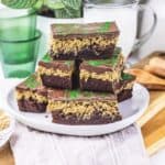 Stacked chocolate oat bars with a green swirl topping on a white plate, inspired by a Dubai Chocolate Brownie Recipe, with glasses and a jug in the background.