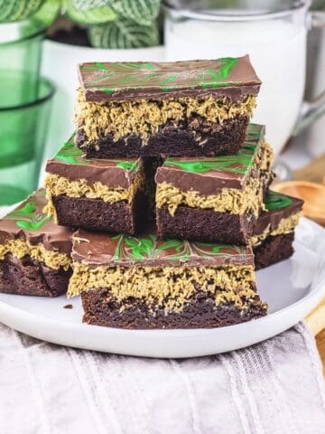 Stacked chocolate oat bars with a green swirl topping on a white plate, inspired by a Dubai Chocolate Brownie Recipe, with glasses and a jug in the background.