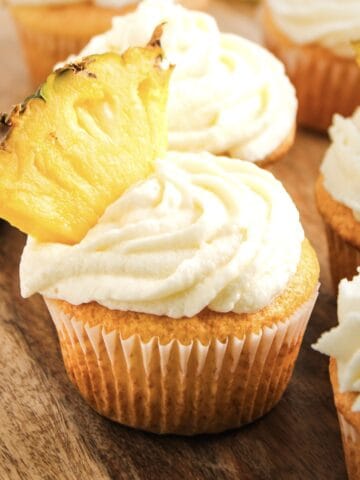 Cupcake with white frosting and a fresh pineapple slice on top, inspired by Dole Whip cupcakes, placed on a wooden surface.