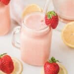 A pink smoothie in a mason jar with a glass straw, garnished with a strawberry, surrounded by lemons and strawberries&mdash;perfect for anyone craving a refreshing Frozen Strawberry Lemonade.