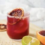 A glass of vibrant blood orange mocktail with ice, garnished with a dried orange slice, sits beside fresh lime and orange halves.