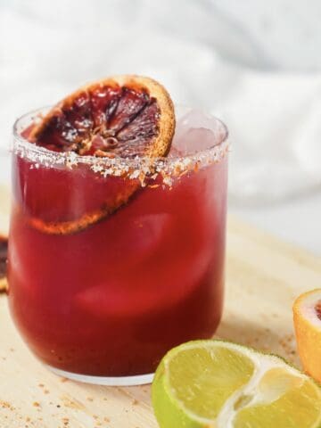 A glass of vibrant blood orange mocktail with ice, garnished with a dried orange slice, sits beside fresh lime and orange halves.