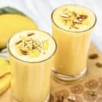Two glasses of Mango Lassi topped with sliced almonds sit on a wooden surface, accompanied by fresh mango and pistachios, showcasing a refreshing Mango Lassi Recipe perfect for any occasion.