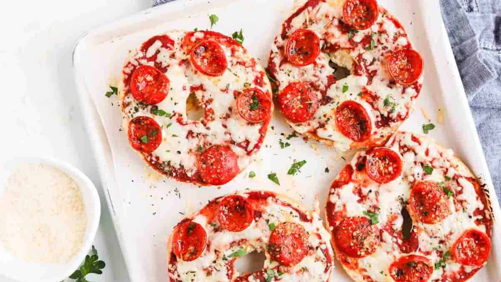 Bagels topped with melted cheese, tomato sauce, and pepperoni, garnished with herbs on a baking tray.