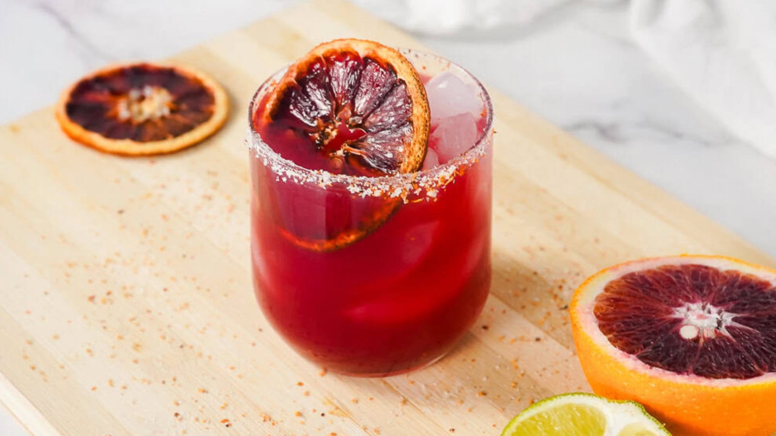A glass of red cocktail with ice, garnished with a blood orange slice, next to lime and orange halves.