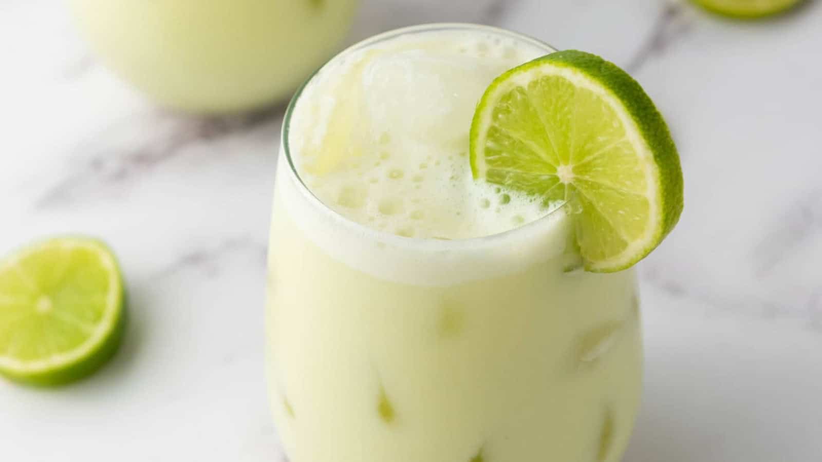 A glass of creamy lime drink with ice, garnished with a lime slice, on a white surface with lime slices nearby.