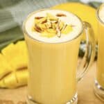 A glass of mango lassi topped with chopped nuts, with mango slices and a green cloth in the background—an inviting scene for any Mango Lassi Recipe lover.