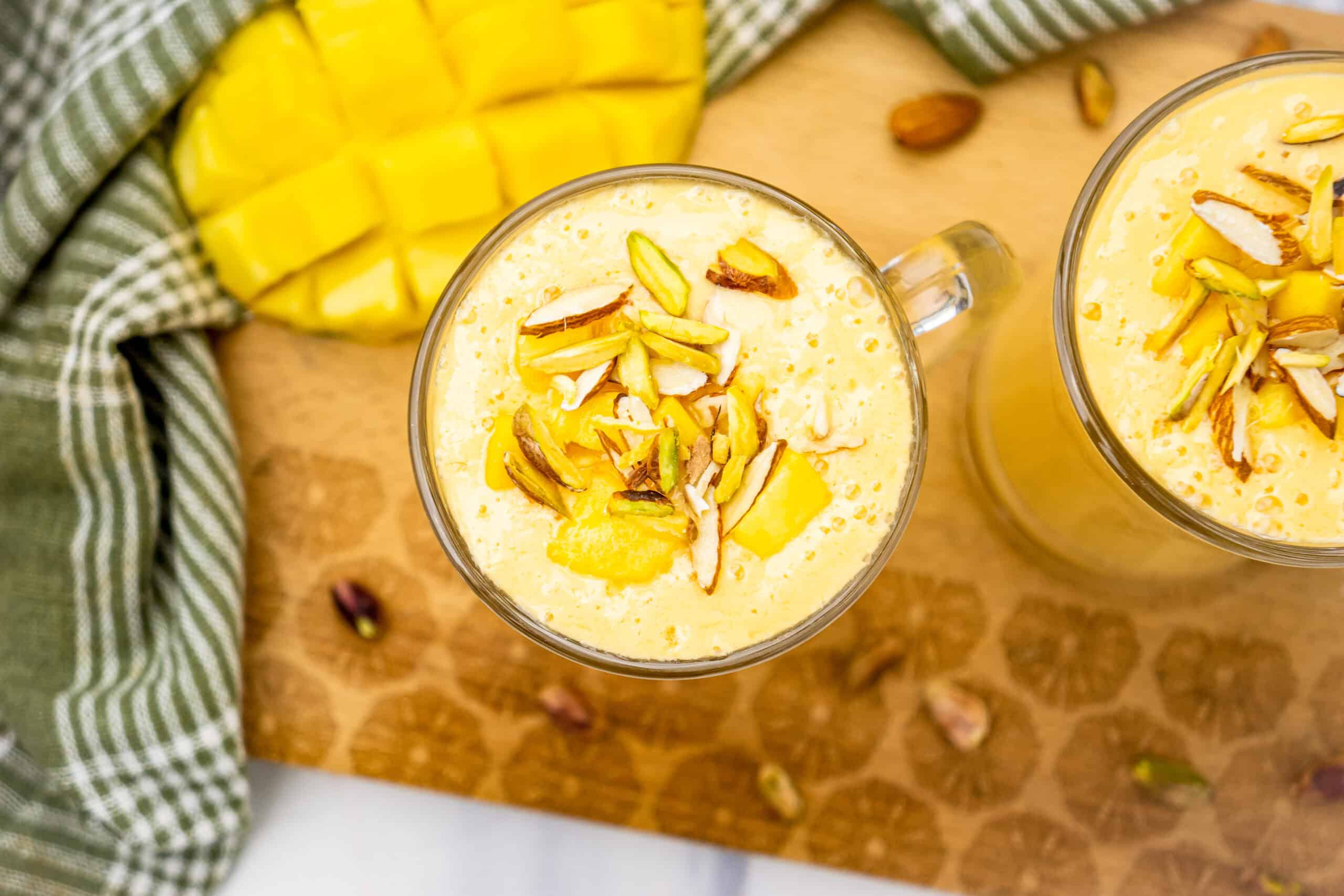 Two glasses of mango lassi, made with our Mango Lassi Recipe and topped with nuts, sit next to diced mango and a green checkered cloth on a wooden board.