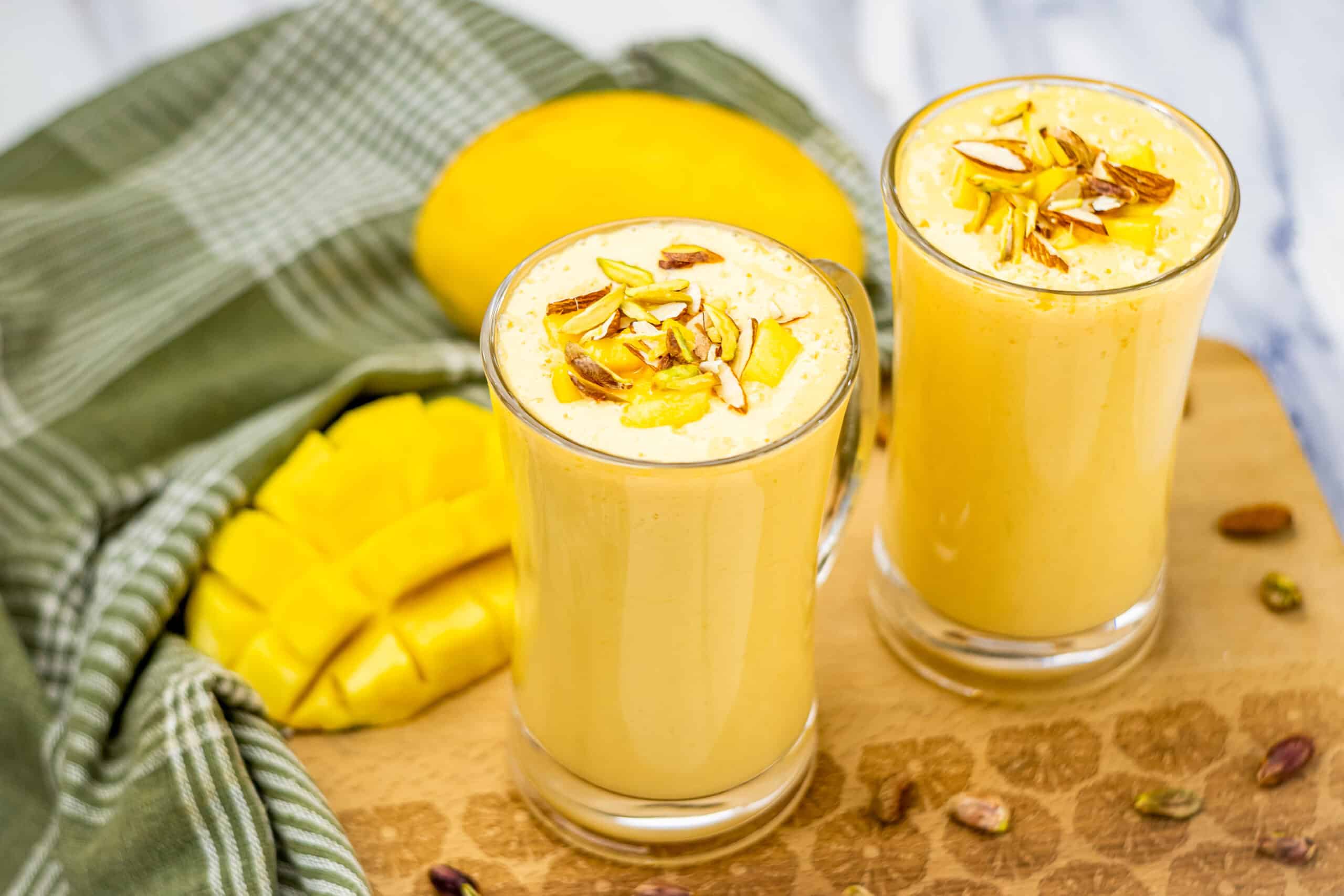 Two glasses of creamy mango lassi, made with our favorite Mango Lassi Recipe, topped with nuts and served beside sliced mango and a green checkered cloth on a wooden board.