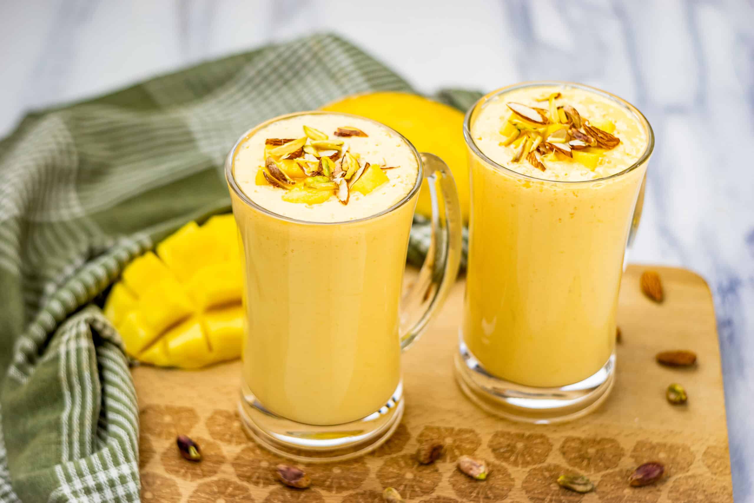 Two glasses of mango lassi topped with sliced nuts, with a ripe mango and green cloth in the background, beautifully showcasing this classic Mango Lassi Recipe on a wooden board.