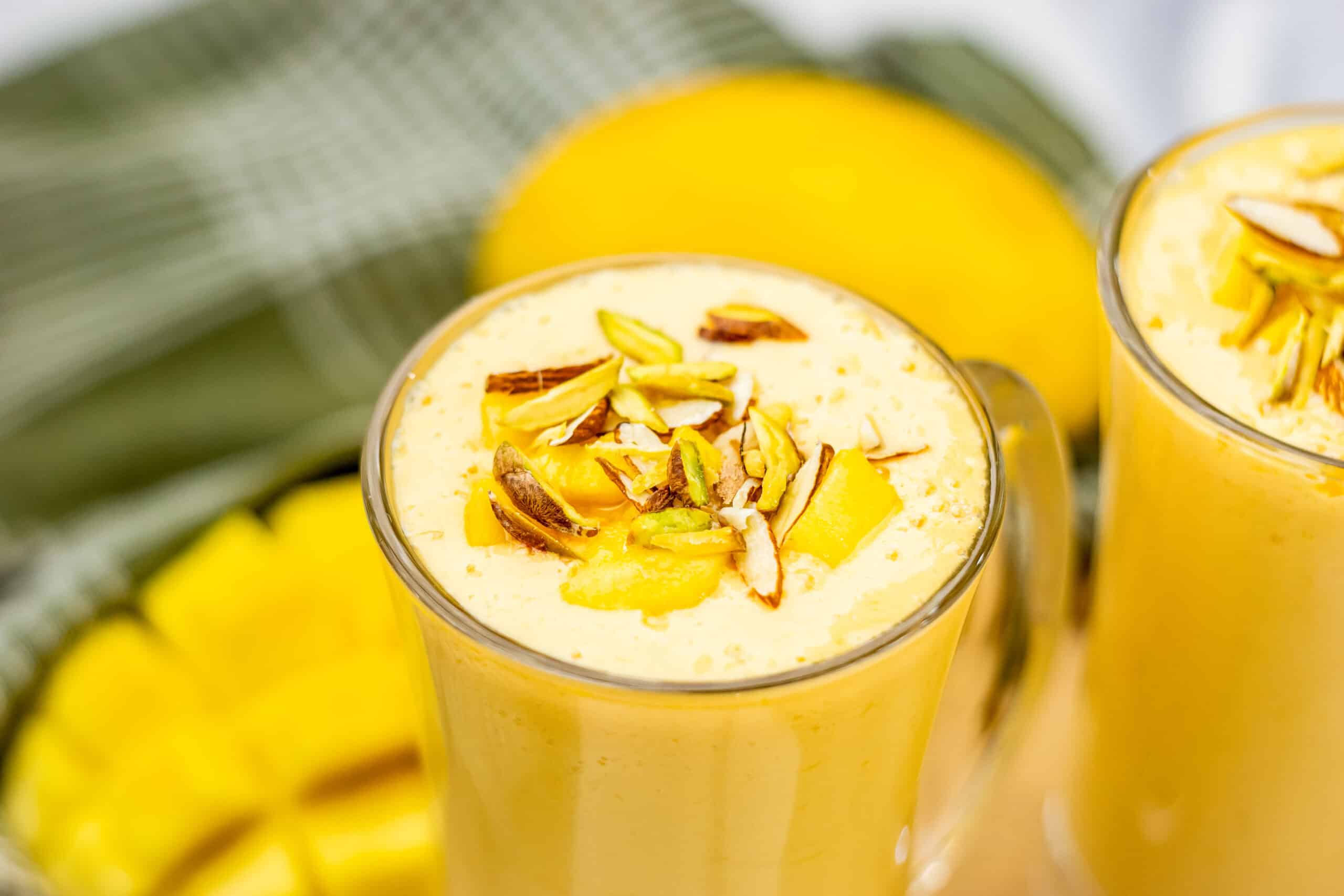 A glass of mango lassi, made from a classic Mango Lassi Recipe, is topped with chopped almonds and mango pieces, with a diced mango in the background.
