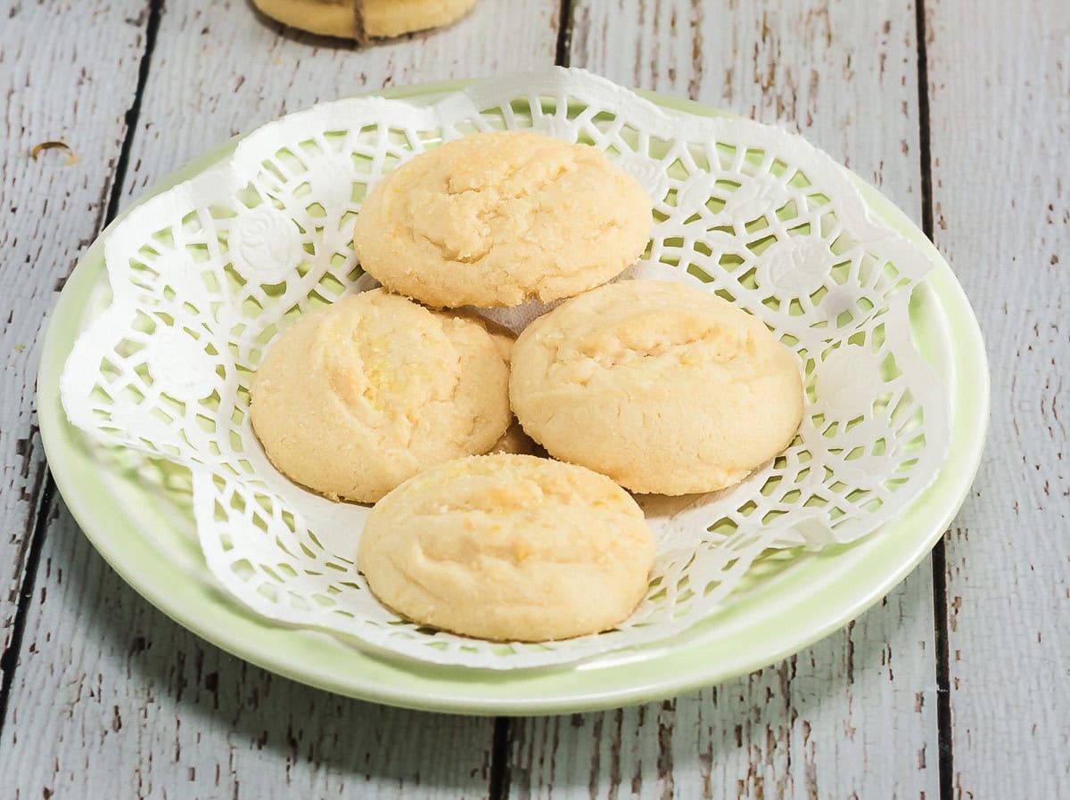 Four round Amish cookies rest on a lace-patterned paper in a green plate atop a wooden surface.