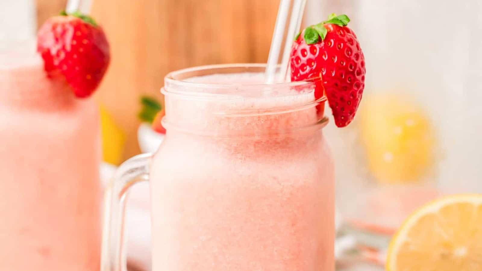 A pink strawberry smoothie in a mason jar with a clear straw and a fresh strawberry on the rim.