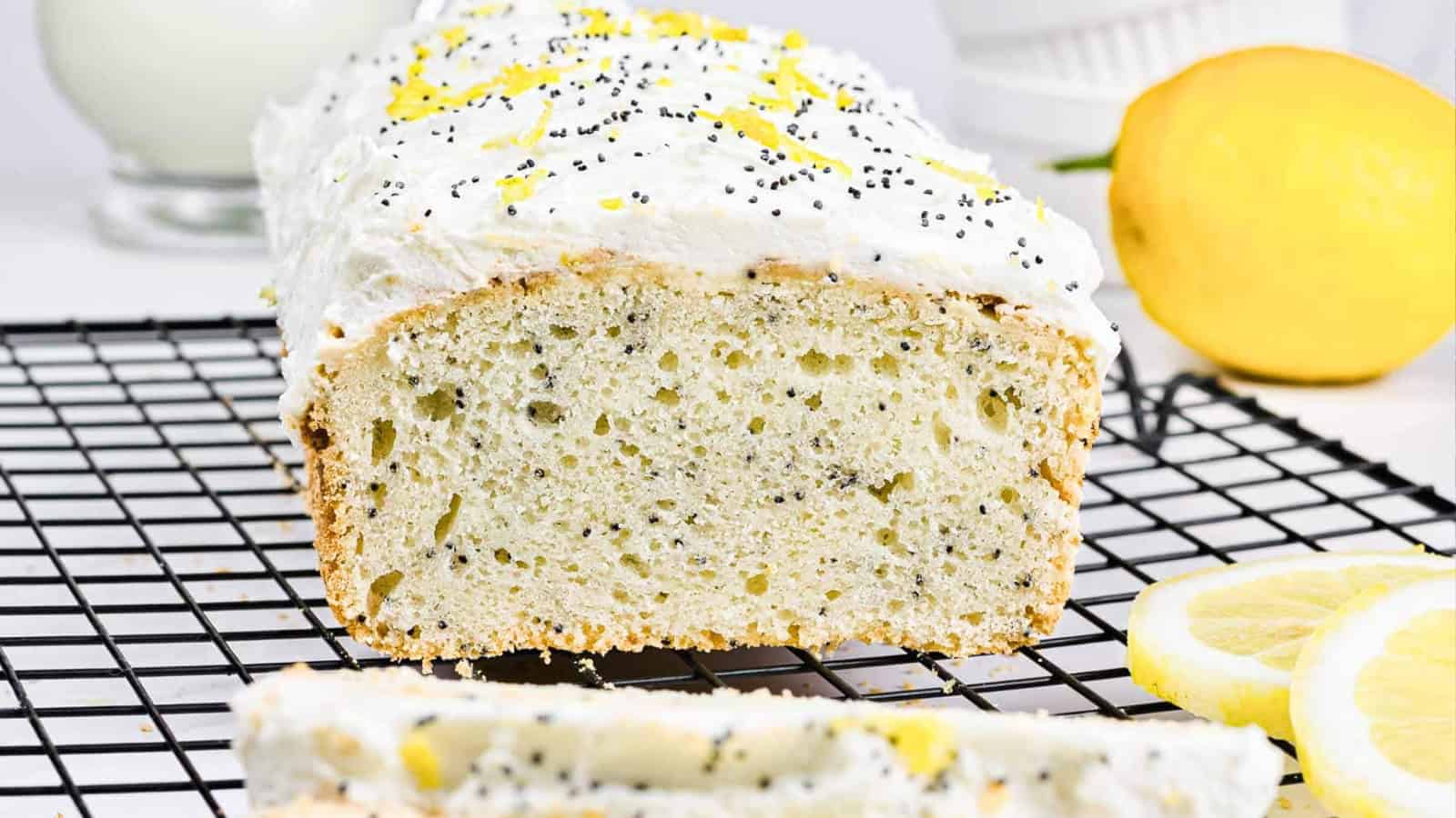 Lemon poppy seed loaf with white frosting, lemon zest, and a sliced lemon on a cooling rack.