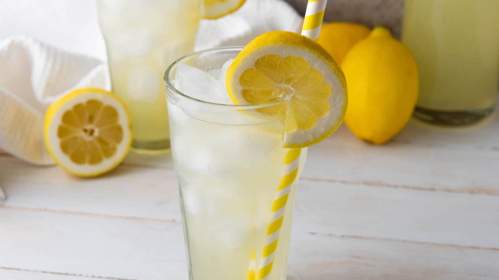 A glass of lemonade with ice, a lemon slice, and a striped straw, with lemons and another glass in the background.