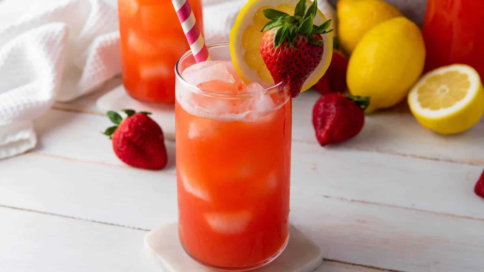 Two glasses of strawberry lemonade with ice, lemon slices, and strawberries, with striped straws on a white table.