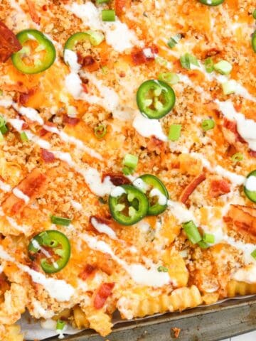 Crinkle-cut fries topped with cheese, bacon, jalapeños, green onions, and drizzled with ranch on a baking sheet.