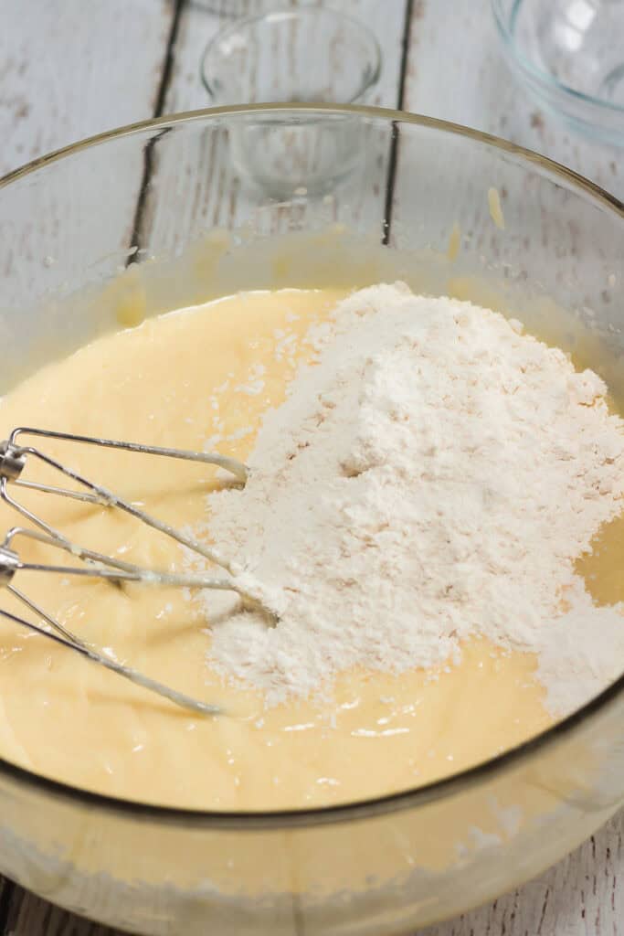 Mixing bowl with yellow cake batter, flour, and metal beaters on a white wooden surface&mdash;perfect for preparing Amish cookies or classic Amish recipes.
