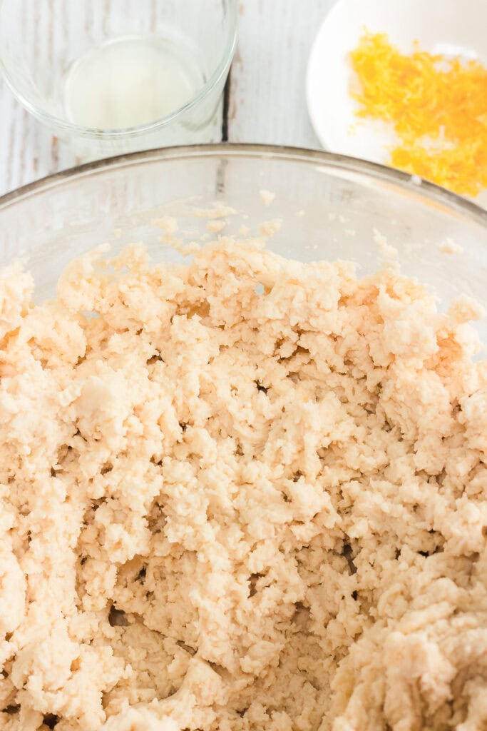 Bowl of cookie dough mixture, ready to become Amish lemon cookies, with lemon zest and juice in small dishes nearby on a white surface.