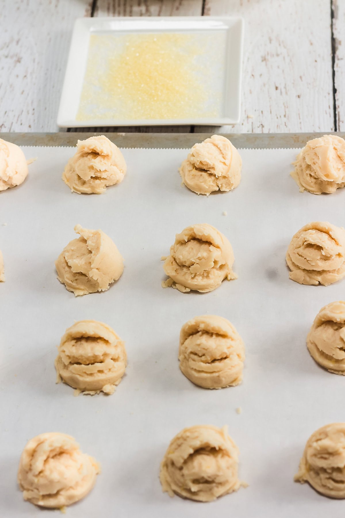 Scoops of Amish cookie dough on a parchment-lined baking sheet with a plate of coarse sugar in the background, ready to become classic Amish cookies.