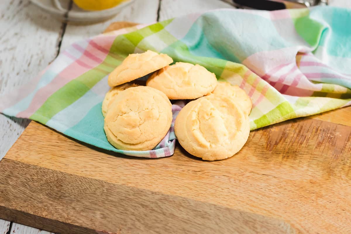 Five Amish cookies rest on a colorful cloth atop a wooden board, with a bowl of fruit in the background.