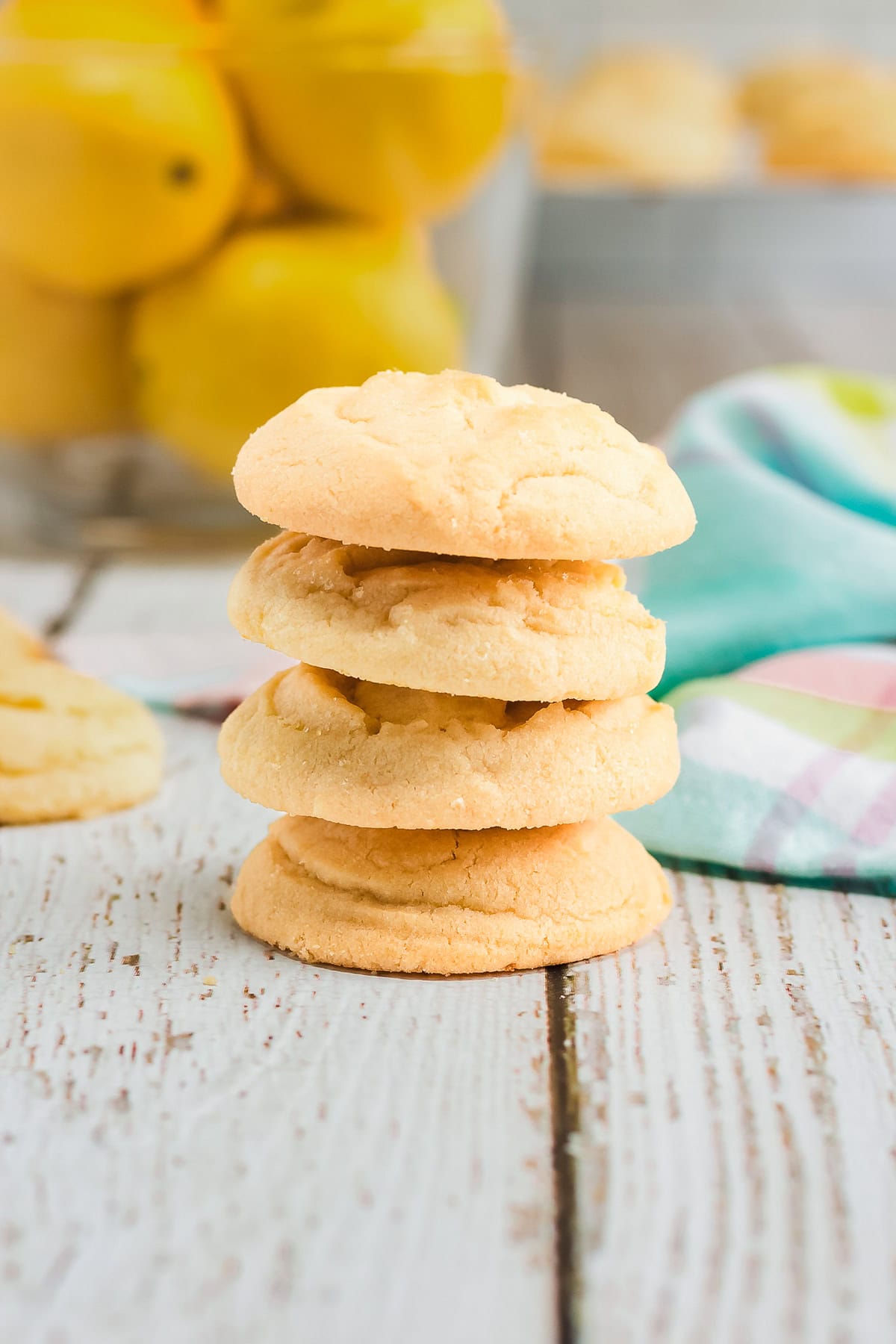 A stack of four golden Amish cookies on a rustic white table with lemons and a colorful cloth in the background.