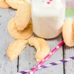 Lemon sugar cookies and a glass bottle of milk with colorful straws rest on a rustic white wooden surface, inspired by classic Amish recipes.
