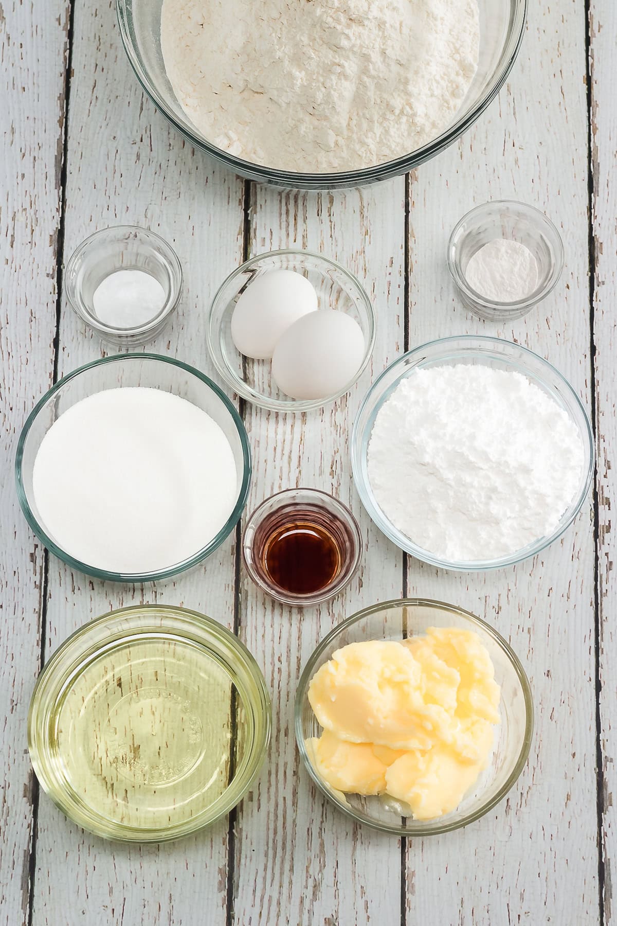 Baking ingredients for Amish cookies: flour, sugar, eggs, butter, oil, vanilla, salt, baking powder, and powdered sugar are arranged on a table.
