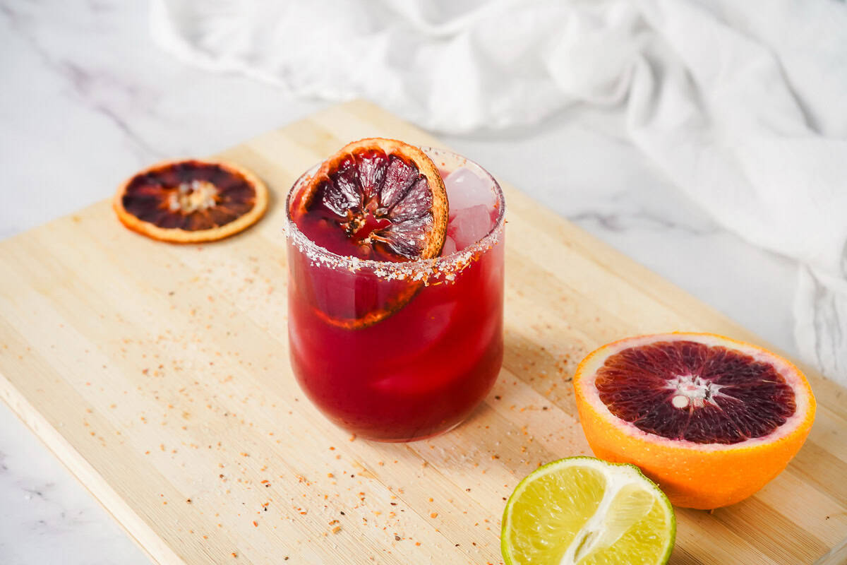 A glass of Blood Orange Margarita Mocktail with ice and a blood orange slice, beside halved blood orange and lime on a wooden board.