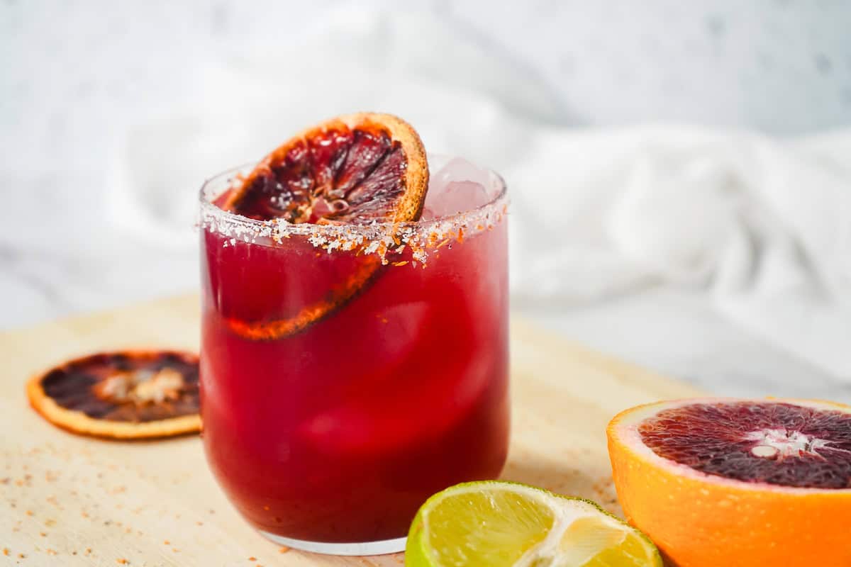 A red Blood Orange Margarita Mocktail with ice, garnished with a blood orange slice, sits beside fresh lime and blood orange halves.